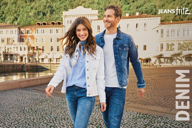 Ein Mann und eine Frau, gekleidet in weißer und blauer Jeans, gehen Hand in Hand über eine kopfsteingepflasterte Straße, vor einer malerischen Kulisse aus weißen Gebäuden und einem klaren blauen Himmel.