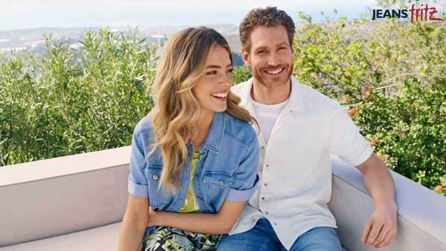 Ein Paar in blauer Jeans und weißer Kleidung sitzt auf einer weißen Couch in einem üppigen Garten, lächelt und umarmt sich.