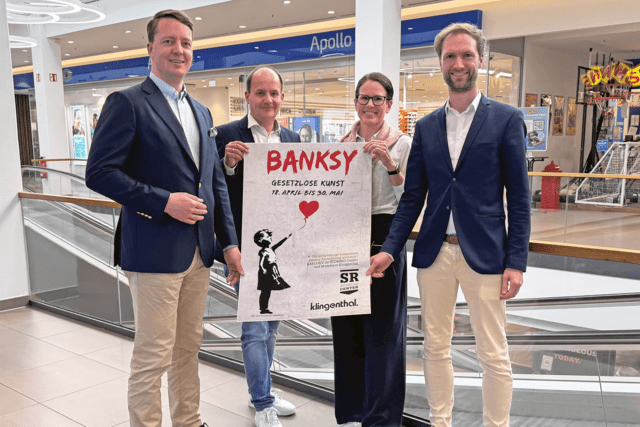 Vier Personen halten in einem Einkaufszentrum ein "BANKSY"-Plakat mit dem Schwarz-Weiß-Bild eines Mädchens mit einem roten Herz, das für eine Wohltätigkeitsveranstaltung wirbt.
