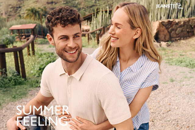 Ein Mann in einem beigen Polohemd und eine Frau in einem blau-weiß gestreiften Hemd umarmen sich in einem üppigen Garten, wobei der Text "JEANS FRITZ" und "SUMMER FEELING" angezeigt wird.